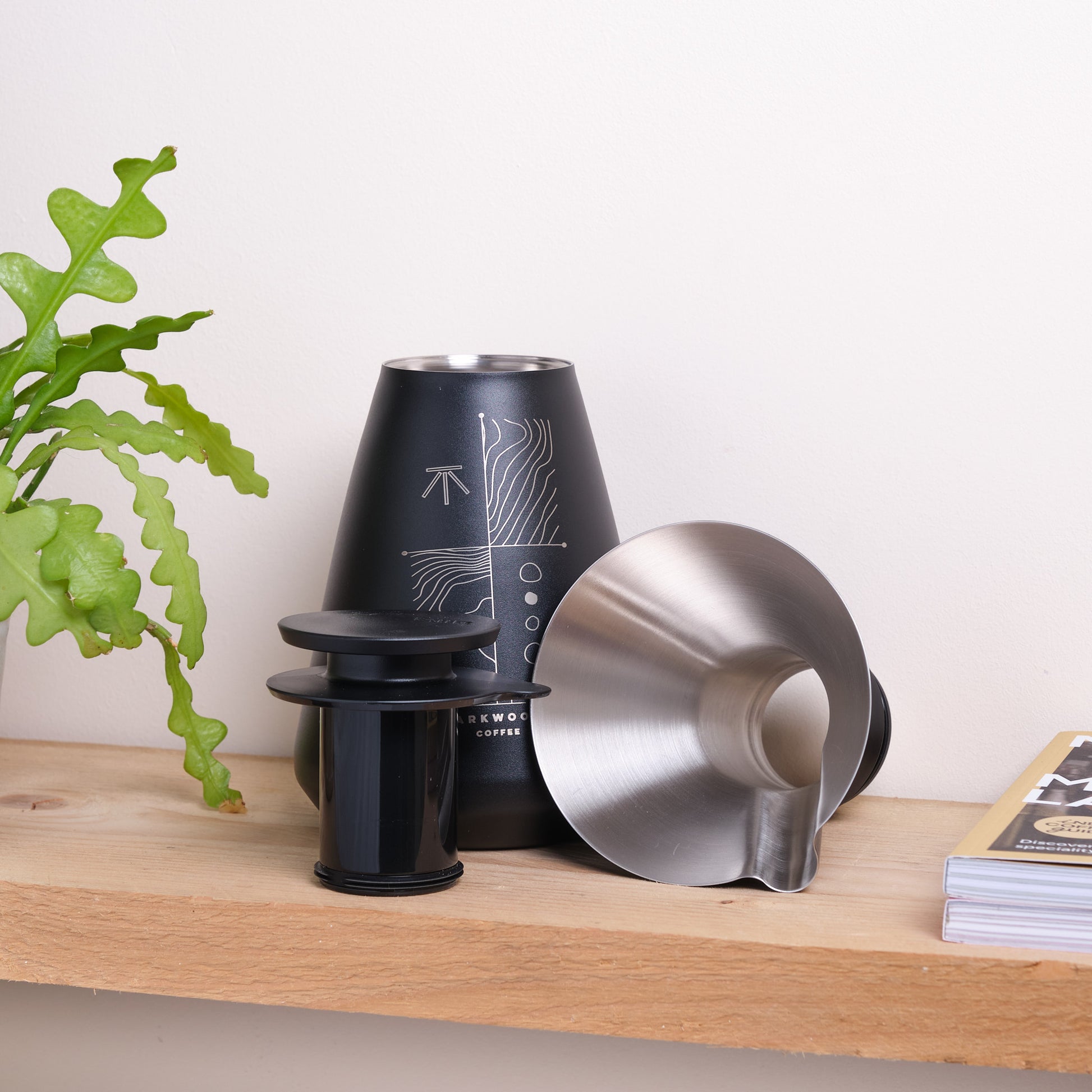 Black coffee brewing flask on a wooden surface with a plant and a magazine in the background