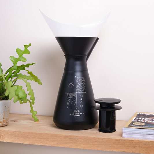 Black coffee brewing flask on a wooden surface with a plant and a magazine in the background