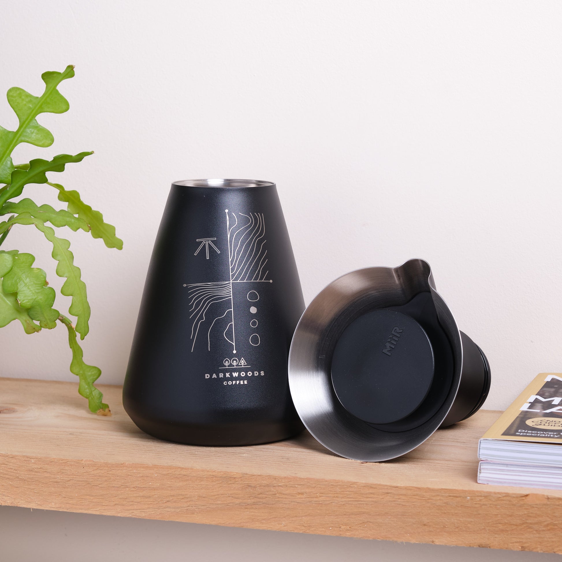 Black coffee brewing flask on a wooden surface with a plant and a magazine in the background